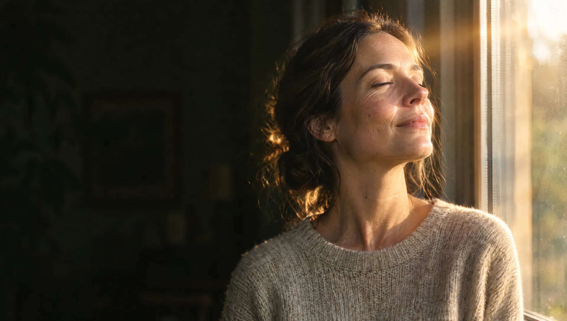 A woman by a window in morning sun with her eyes closed, taking a slow breath.